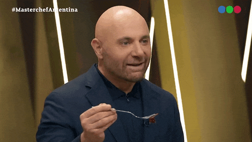 a bald man in a suit is holding a fork in front of a screen that says masterchefargentina