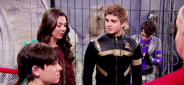 a group of people standing in front of a fence with one of them wearing a black and gold jacket with the letter t on it