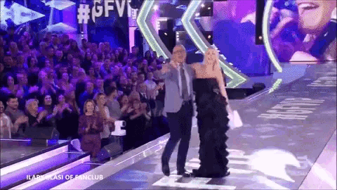 a woman in a black dress is walking down a runway with a man in a suit behind her