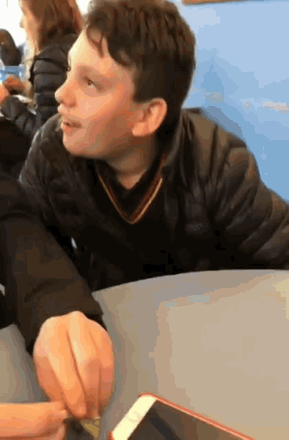 a boy in a black jacket sits at a table with a cell phone in front of him