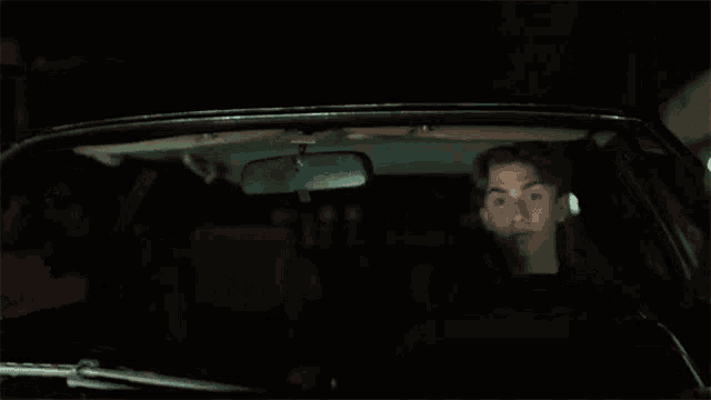 a young man is driving a car at night and looking in the rear view mirror .