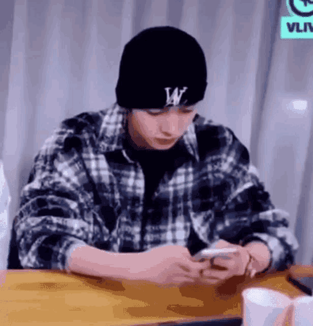 a young man wearing a plaid shirt and a black beanie is sitting at a table looking at his cell phone .