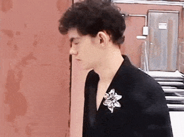 a man with curly hair is wearing a black jacket with a flower brooch on his collar .