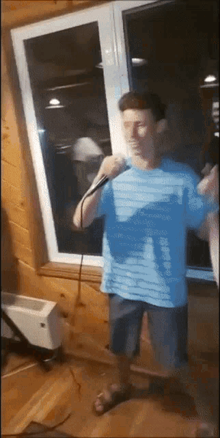 a man in a blue shirt is singing into a microphone in front of a window