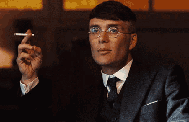 a man wearing glasses and a suit holds a cigarette in his right hand