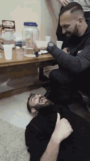 a man laying on the floor giving a thumbs up while another man kneels next to him