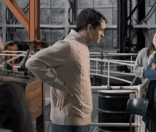 a man in a tan sweater stands with his hands on his back