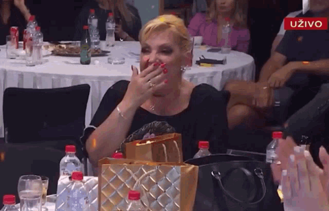 a woman covering her mouth with her hand while sitting at a table with bottles of water and a sign that says uzivo