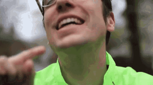 a man wearing glasses and a neon green jacket is laughing and pointing at his face .