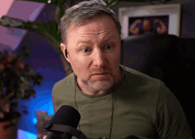 a man wearing ear buds and a green shirt stands in front of a microphone and looks surprised