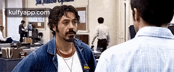 a man with a beard and a denim jacket is talking to another man in a room .