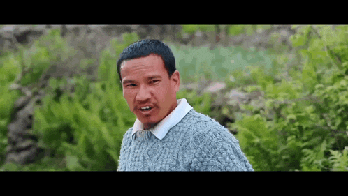 a man in a gray sweater and white shirt is standing in front of a bush .