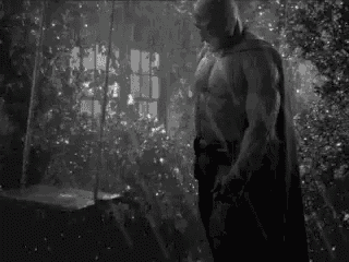 a black and white photo of a man in a batman costume standing in a forest .