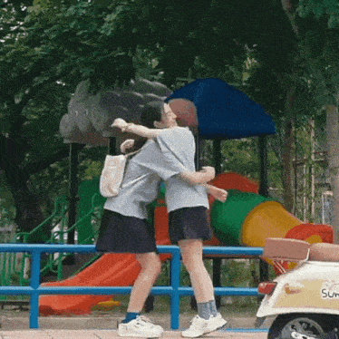 two girls hugging in front of a playground and a yellow scooter with the word sun on the back
