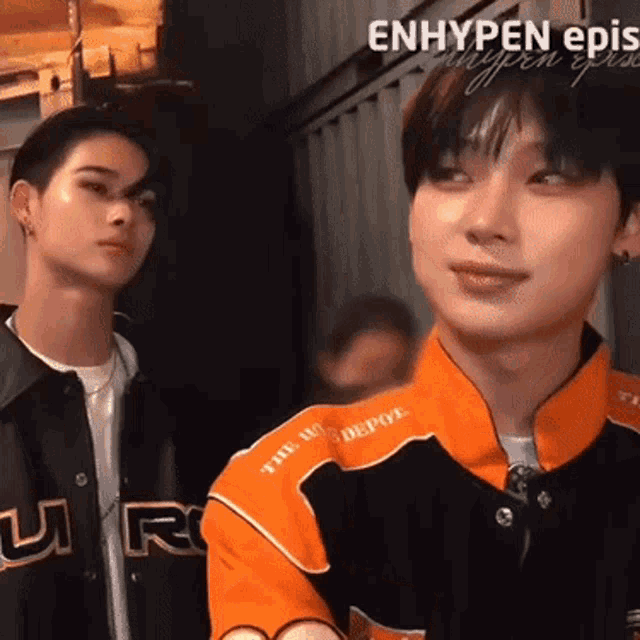 two young men are standing next to each other in a room and one of them is wearing an orange jacket .