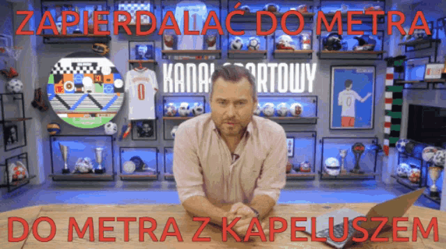 a man sits at a table in front of a wall with soccer balls and a sign that says zapierdalac do metra