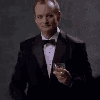 a man in a tuxedo and bow tie is holding a glass of whiskey and pointing .