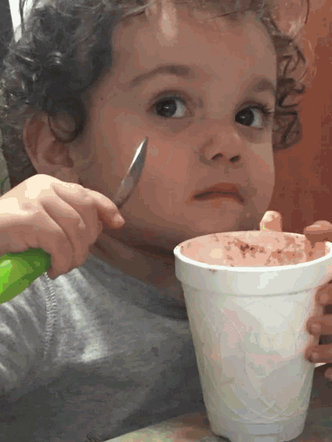 a little girl with curly hair is eating from a styrofoam cup while holding a knife