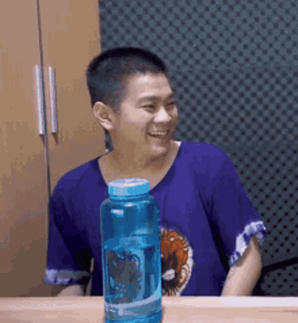 a man in a purple shirt is sitting at a table with a bottle of water .