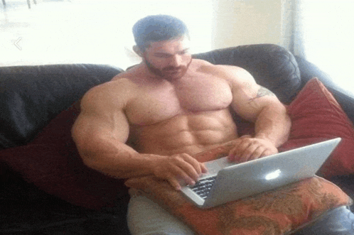 a shirtless man sits on a couch using a laptop