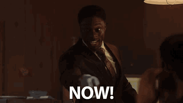 a man in a suit and tie is pointing at the camera with the word now written below him
