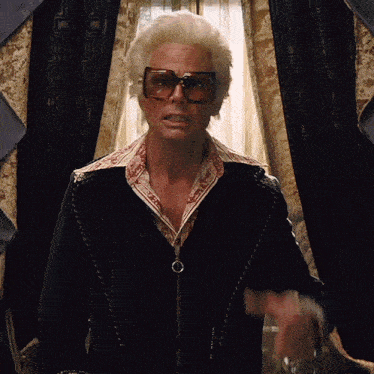 a man wearing sunglasses and a black jacket stands in front of curtains