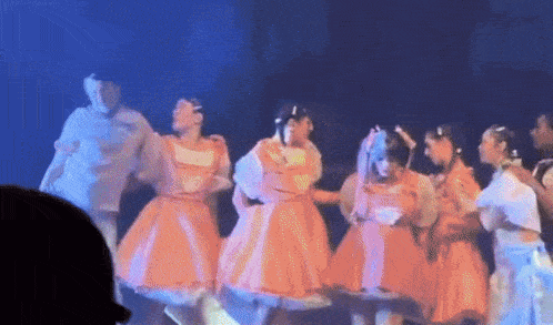 a group of young girls in orange dresses are dancing on a stage .