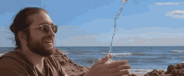 a man wearing sunglasses is sitting on a beach with water pouring into his hand