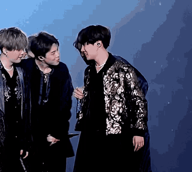 a man in a sequined jacket holds a microphone and talks to two other men