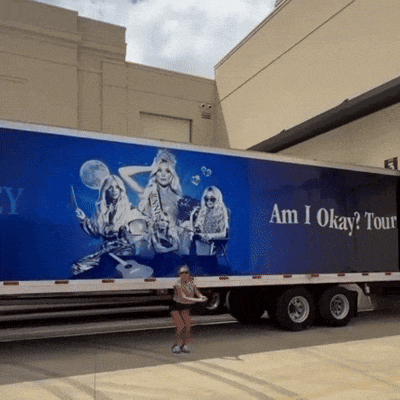 a woman stands in front of a large blue truck that says am i okay tour