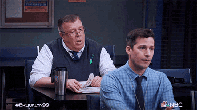 two men sitting at a table with a nbc logo on the bottom right