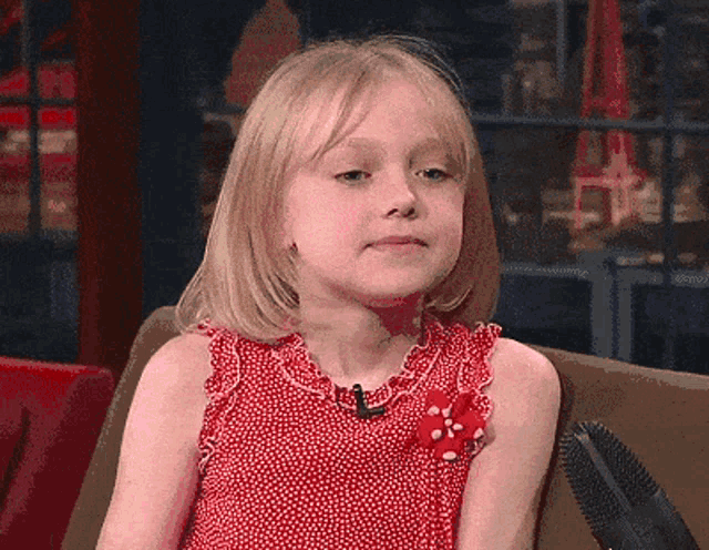 a little girl in a red dress sits on a couch with a microphone