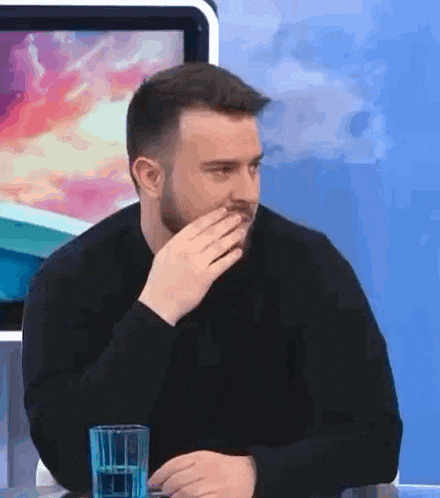 a man with a beard is sitting at a table with a glass of water and covering his mouth with his hand .