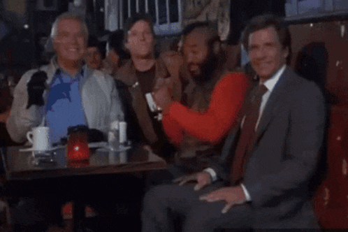 a group of men are sitting at a table and one of them is wearing a red shirt with the letter m on it