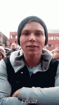 a man wearing a beanie and a vest is talking to a group of people .
