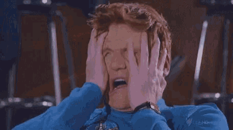 a man in a blue sweatshirt is covering his face with his hands and making a funny face .