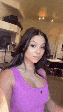a woman in a purple tank top is standing in a living room looking at the camera .