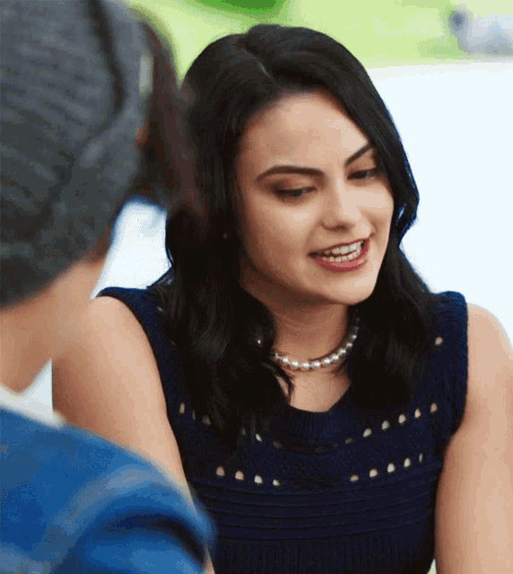 a woman wearing a pearl necklace smiles while sitting next to a man wearing a beanie
