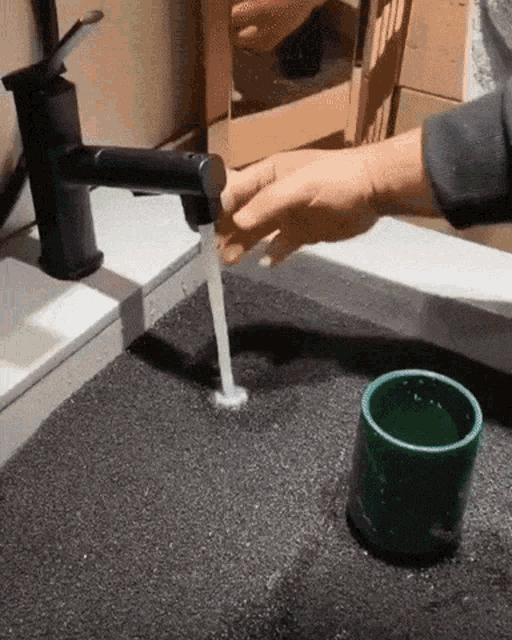 a person is pouring water from a black faucet into a green cup .