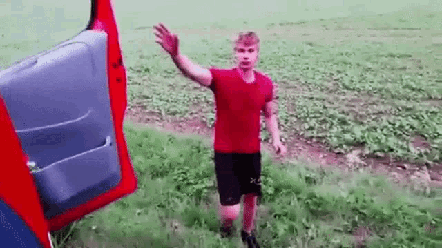 a man in a red shirt and black shorts is standing in a field with his hand up .