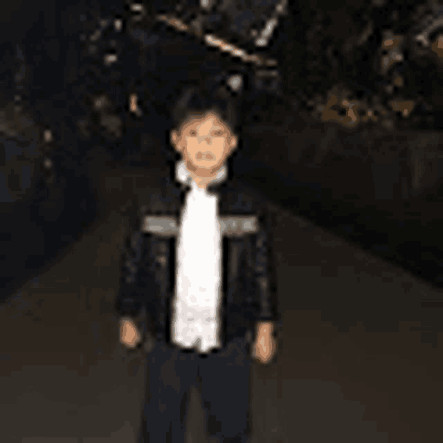 a young boy wearing a leather jacket and a white shirt is standing on a sidewalk .