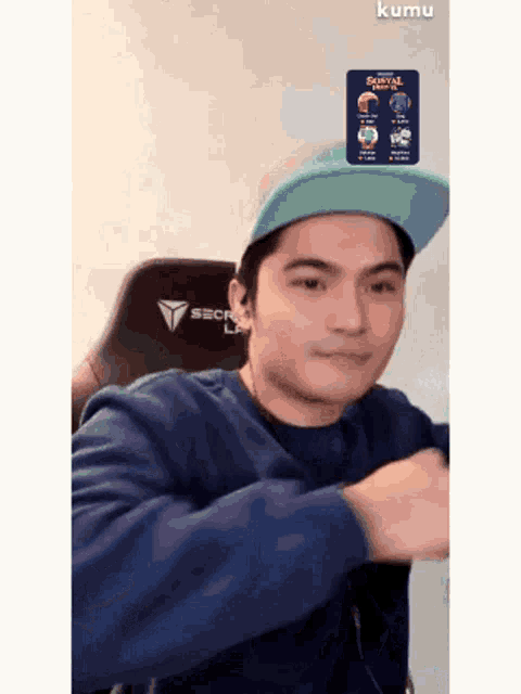 a man wearing a blue shirt and a hat is sitting in a chair with a sticker on his head that says kumu