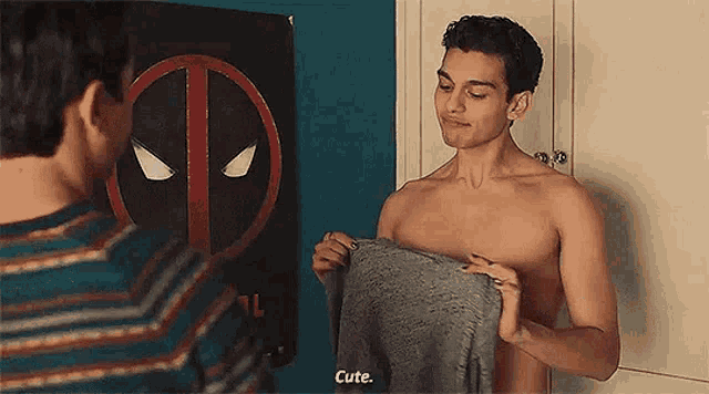 a shirtless man is holding a sweater and talking to another shirtless man in front of a deadpool poster .