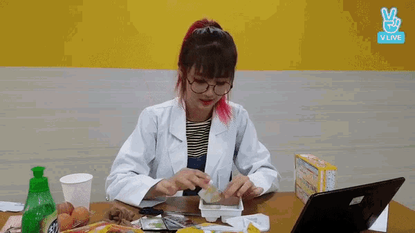 a woman wearing glasses and a lab coat is sitting at a table with a laptop and food .