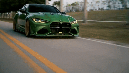 a green bmw is driving down a road with trees in the background .