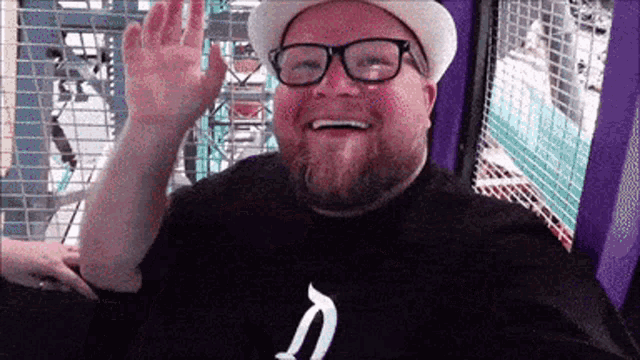a bearded man wearing glasses and a hat is waving