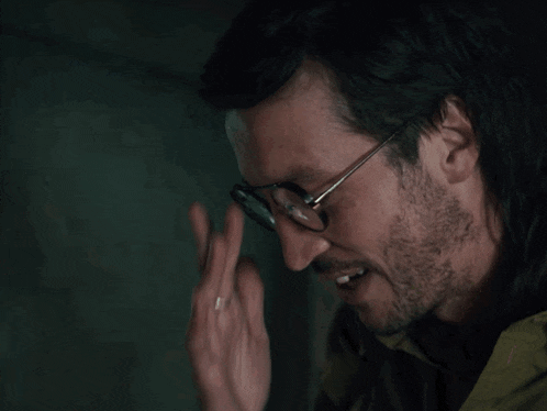 a man wearing glasses and a green jacket holds his hand to his head