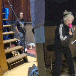 a woman singing into a pink microphone with a man standing behind her