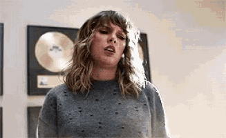 taylor swift is wearing a gray sweater and standing in front of a wall with plaques on it .