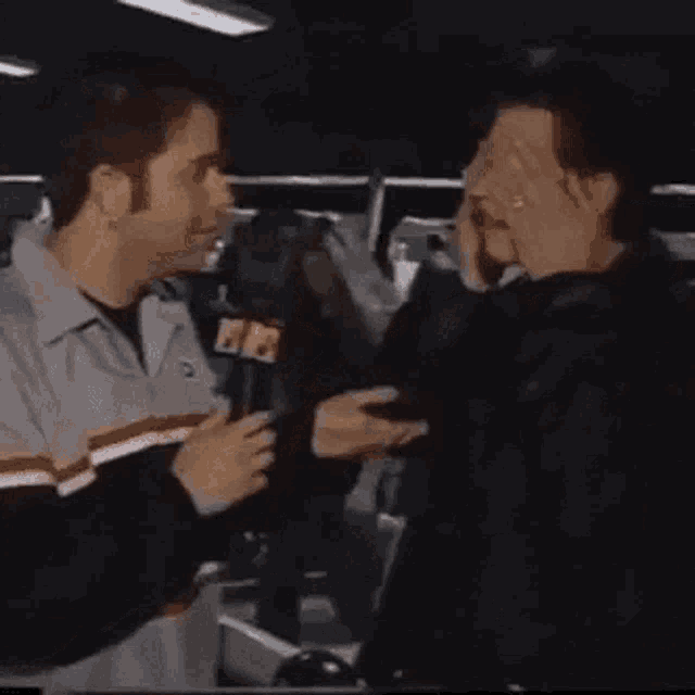 two men are talking to each other in a parking lot while holding a microphone .